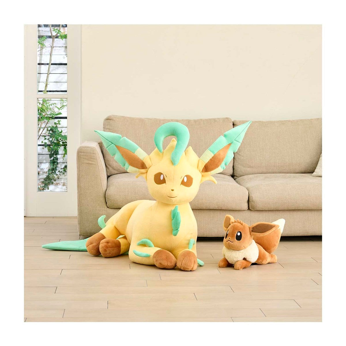 A large yellow and mint green Eevee plush toy sits on a light wood floor in front of a beige sofa, with a smaller brown and cream Eevee plush toy beside it.