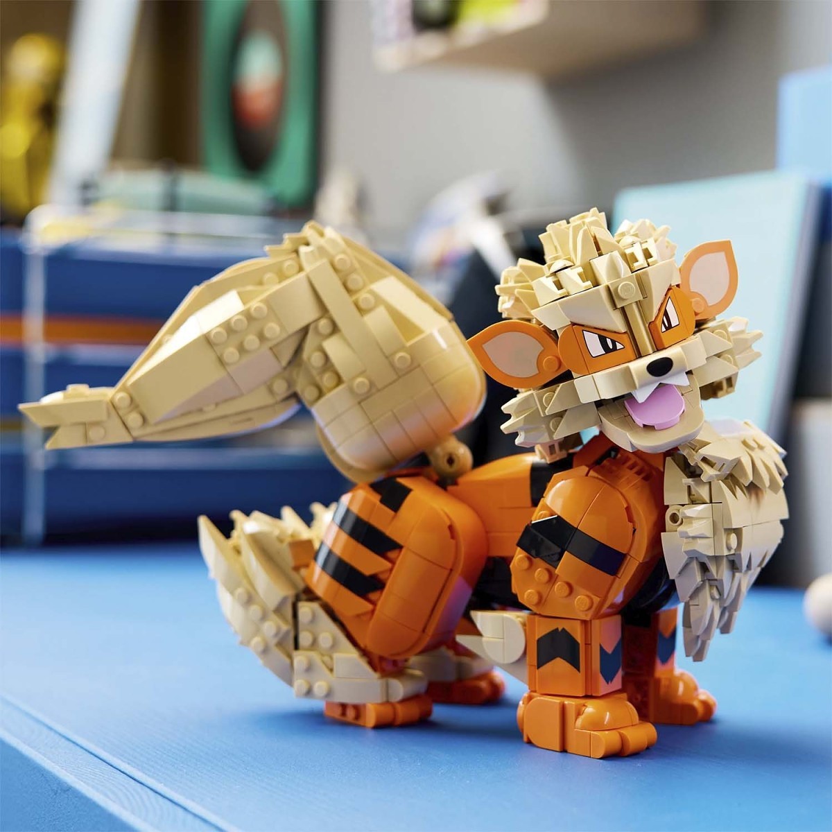 A toy building-block animal character resembling a fox or tiger, assembled from orange and beige bricks, standing on a blue surface with a blurred indoor background.