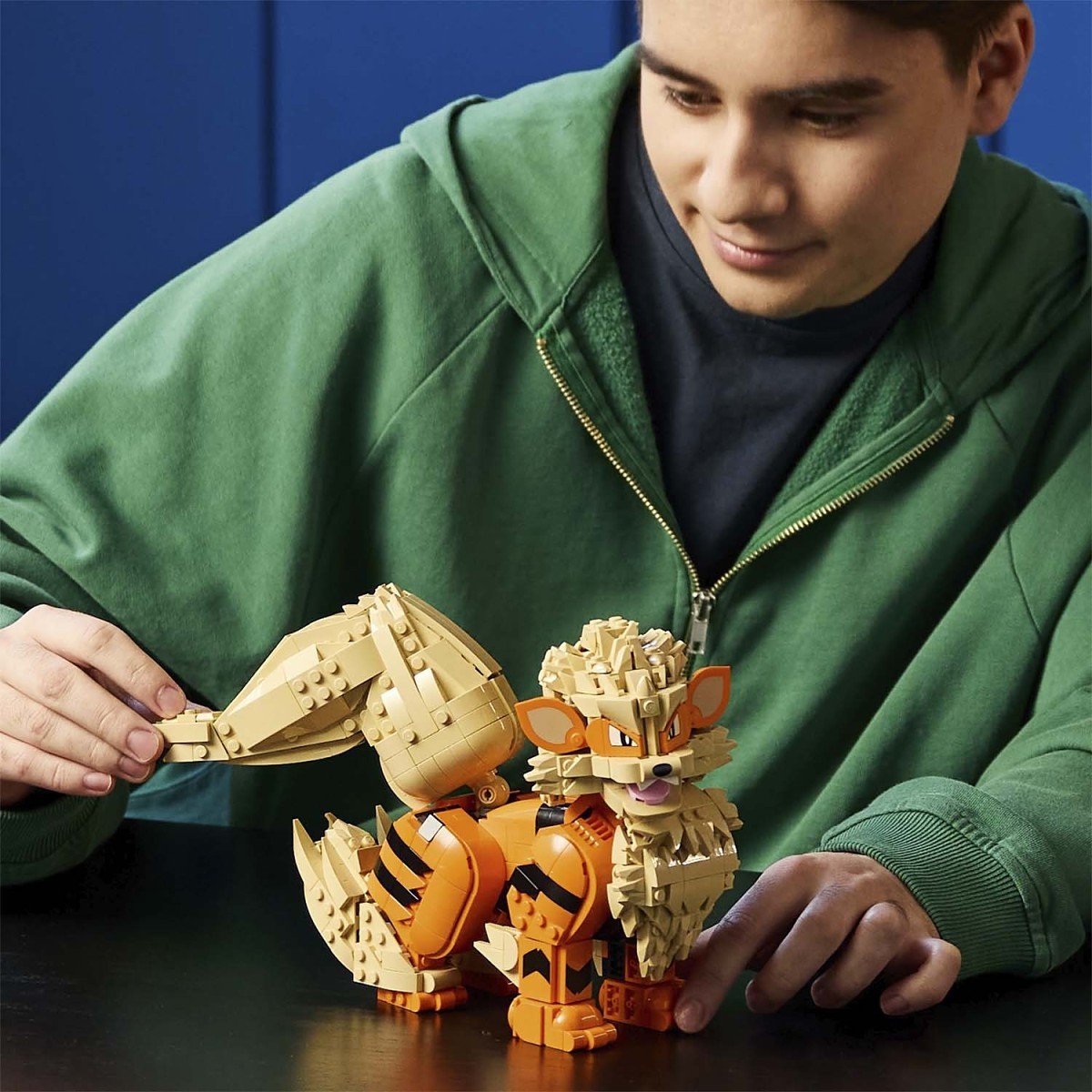 A person in a green hoodie plays with a toy fox built from beige and orange blocks on a dark table against a blue background.