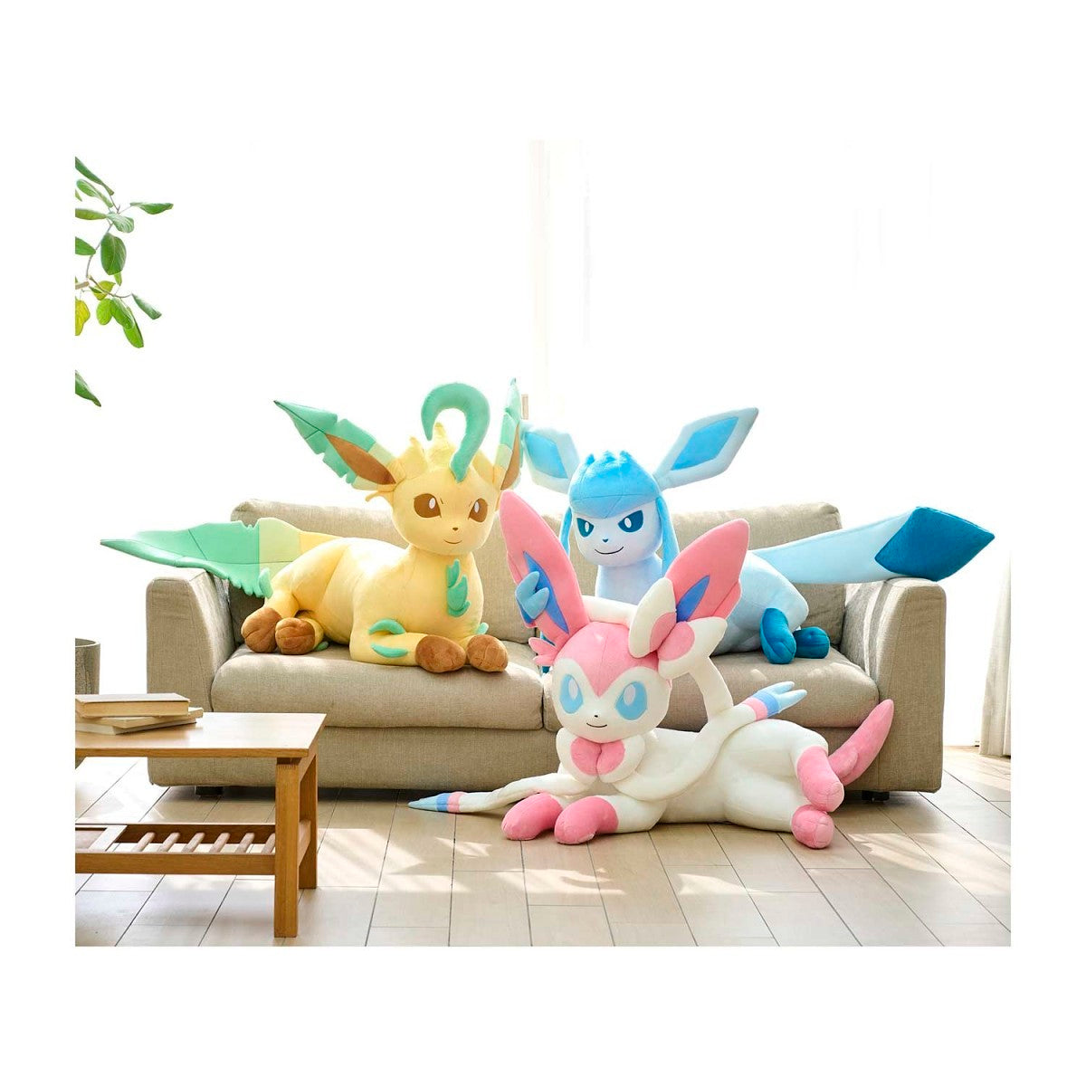 Three large plush toy Pokémon (Eevee, Gliscor, and Mew) arranged on and around a beige sofa in a bright, modern living room.
