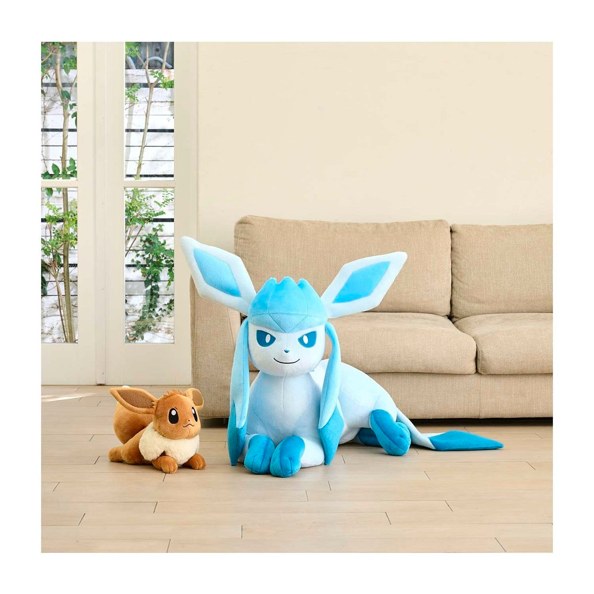 A large Gliscor plush toy in light blue and white is sitting on a light wood floor in front of a beige sofa, with a smaller brown and cream plush toy beside it. The room has a clean, minimalist interior with a window in the background.