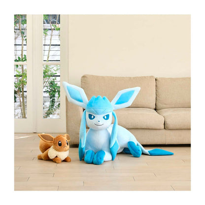 A large Gliscor plush toy in light blue and white is sitting on a light wood floor in front of a beige sofa, with a smaller brown and cream plush toy beside it. The room has a clean, minimalist interior with a window in the background.
