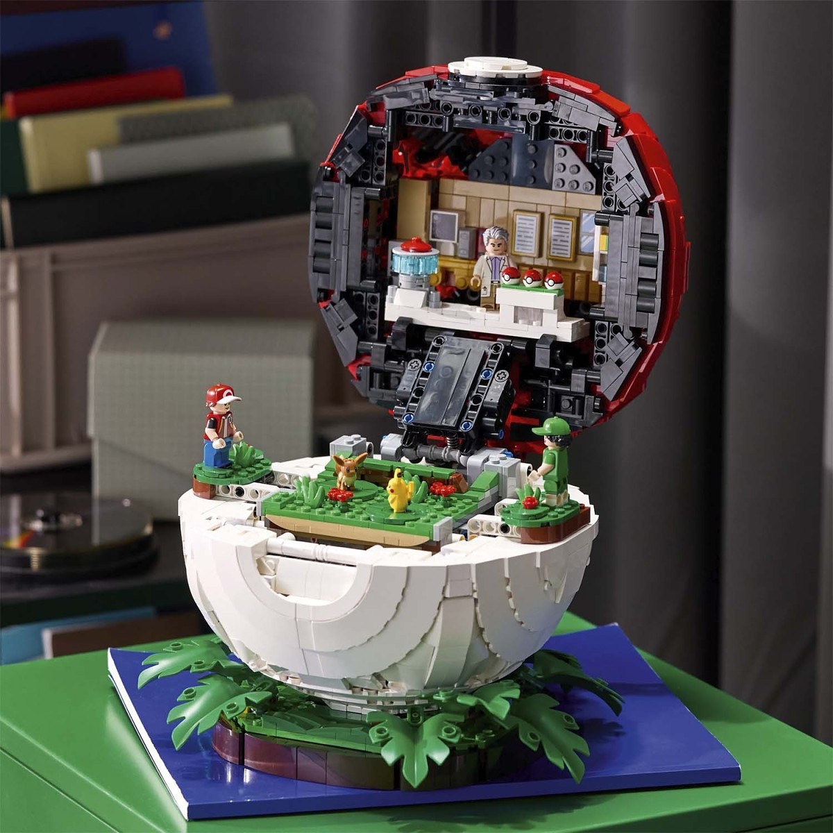 A detailed LEGO-style miniature model of a spherical structure with an open top, featuring green grassy platforms, small character figures, and a furnished interior visible inside the shell. The model is displayed on a blue base against an indoor room background.
