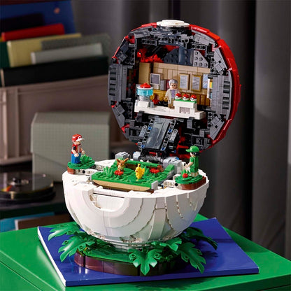 A detailed LEGO-style miniature model of a spherical structure with an open top, featuring green grassy platforms, small character figures, and a furnished interior visible inside the shell. The model is displayed on a blue base against an indoor room background.